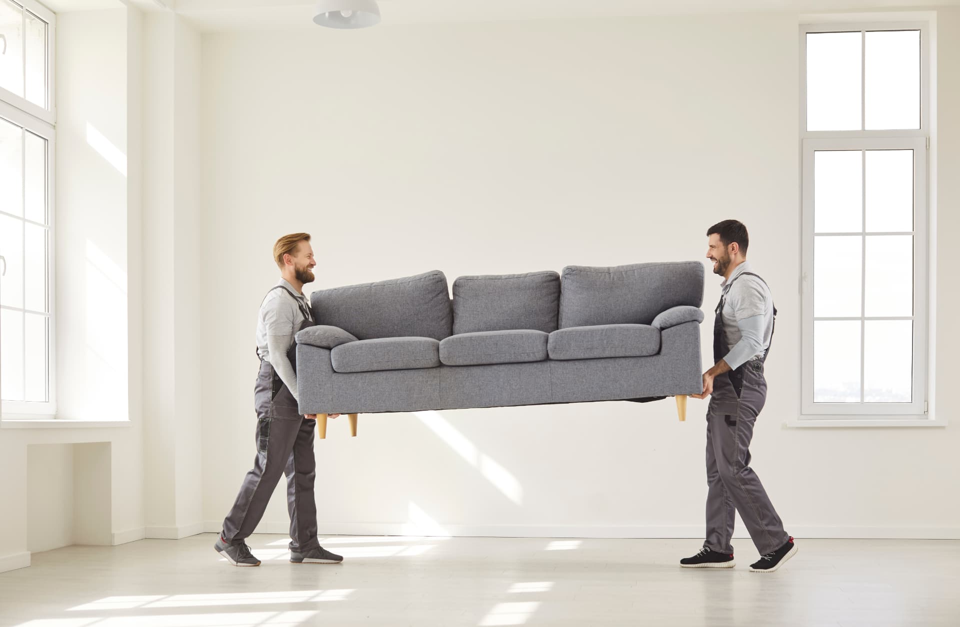 Two delivery fitters carrying a grey fabric sofa into a bright empty living room